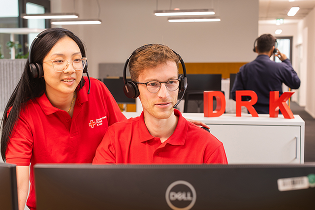 Dieses Bild zeigt zwei Mitarbeitende des DRK in DRK-Poloshirts mit Headsets auf dem Kopf, die gemeinsam auf einen Monitor gucken.
